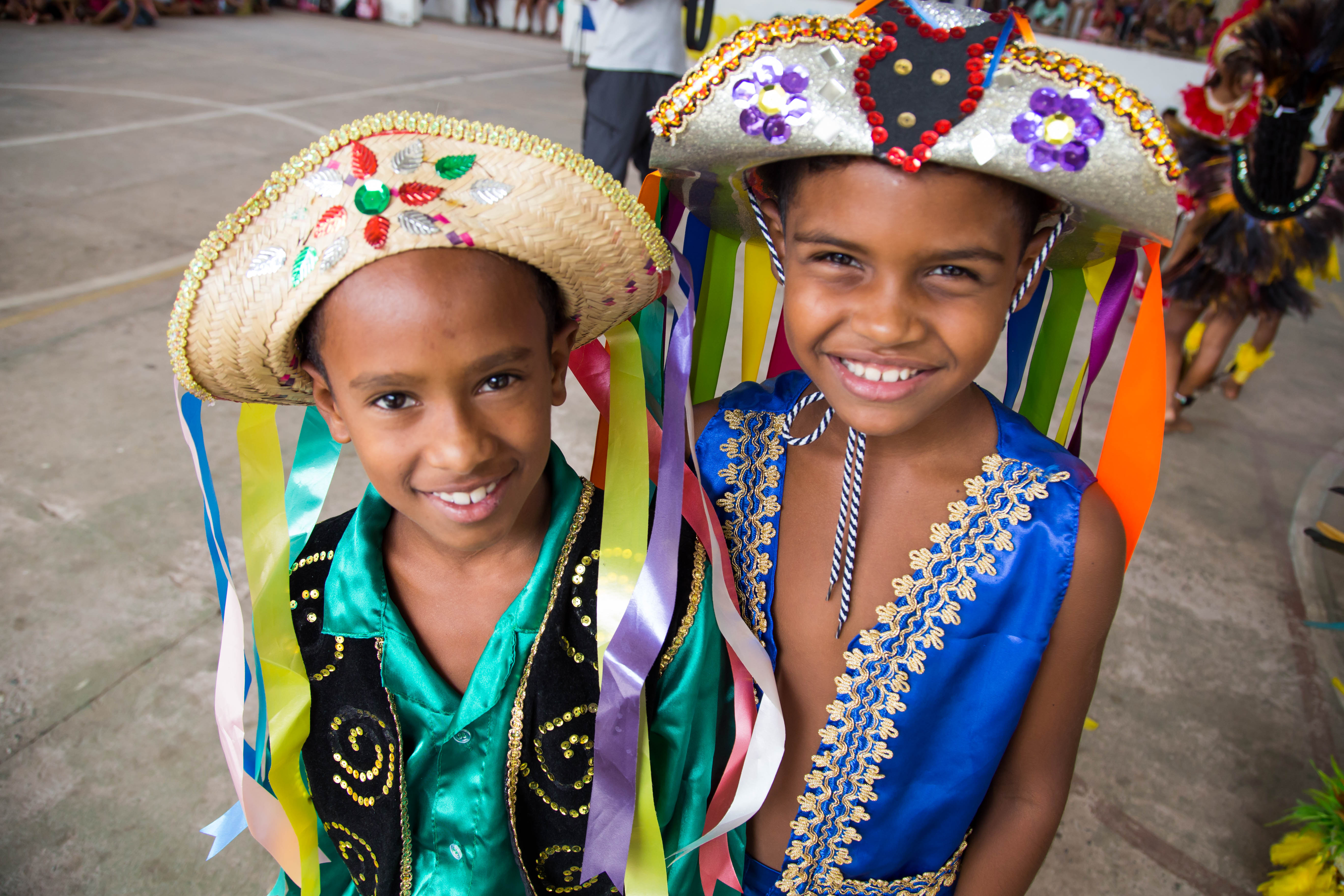 Dois garotos sorridentes posam para foto vestindo chapéus e roupas coloridas.