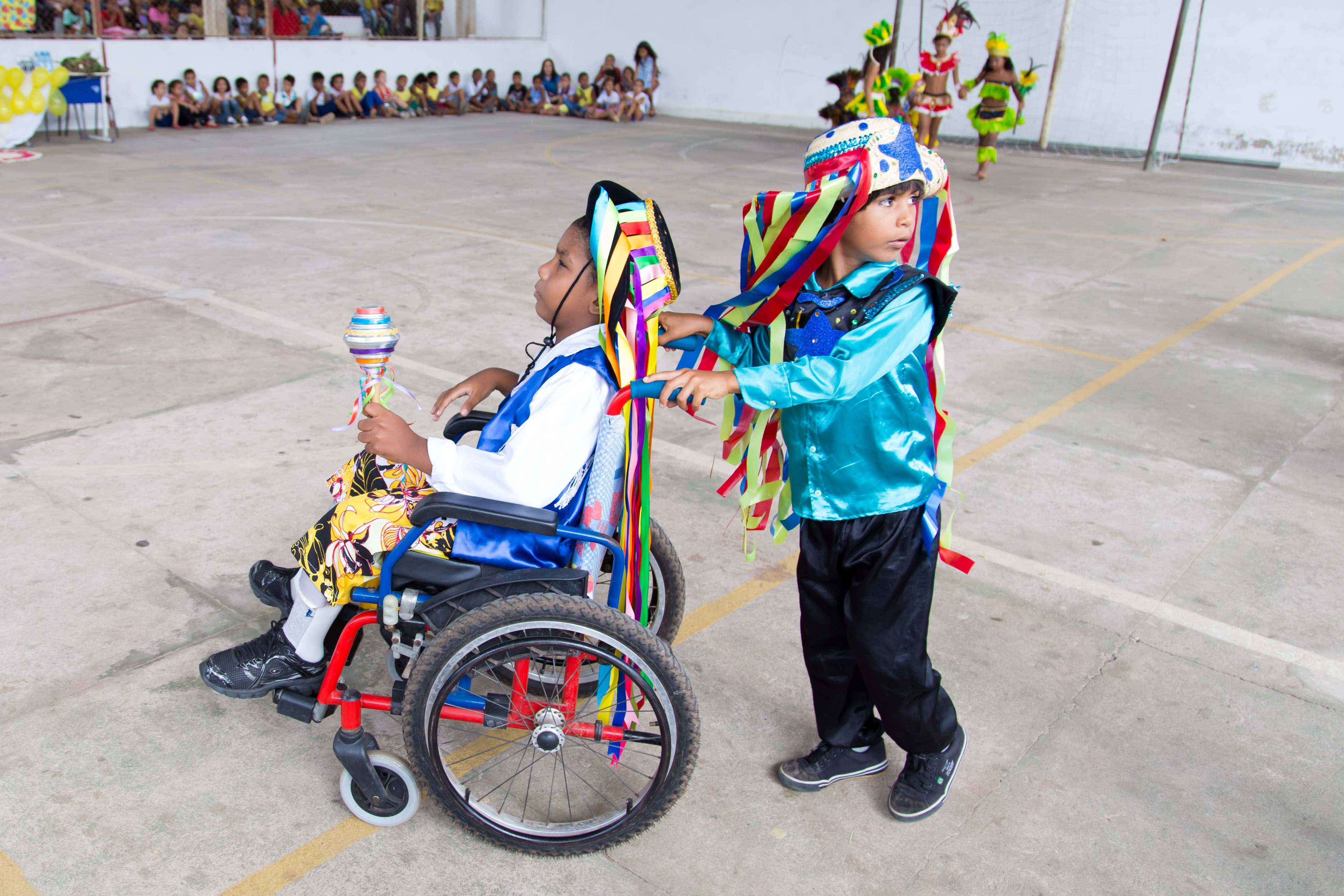 Dois garotos com roupas coloridas dançam. Um deles usa uma cadeira de rodas e o outro o empurra.