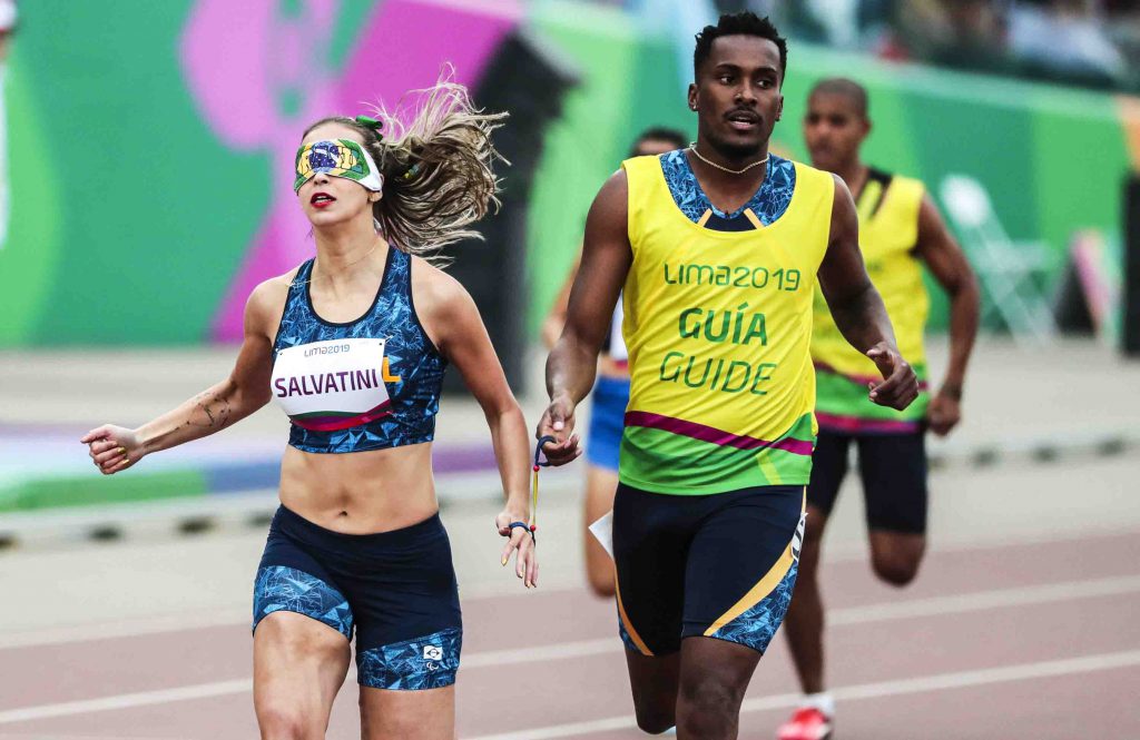 Atleta Lorena Spoladore e seu guia correm na pista dos Jogos Parapanamericanos Lima 2019. A atleta usa uma proteção nos olhos. Fim da descrição.