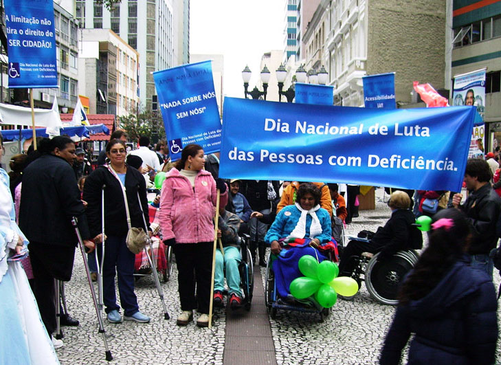 Grupo de pessoas com deficiência realiza passeata em rua de Curitiba em 2008. Duas pessoas seguram um cartaz azul com o seguinte texto: "Dia Nacional de Luta das Pessoas com Deficiência". Fim da descrição.