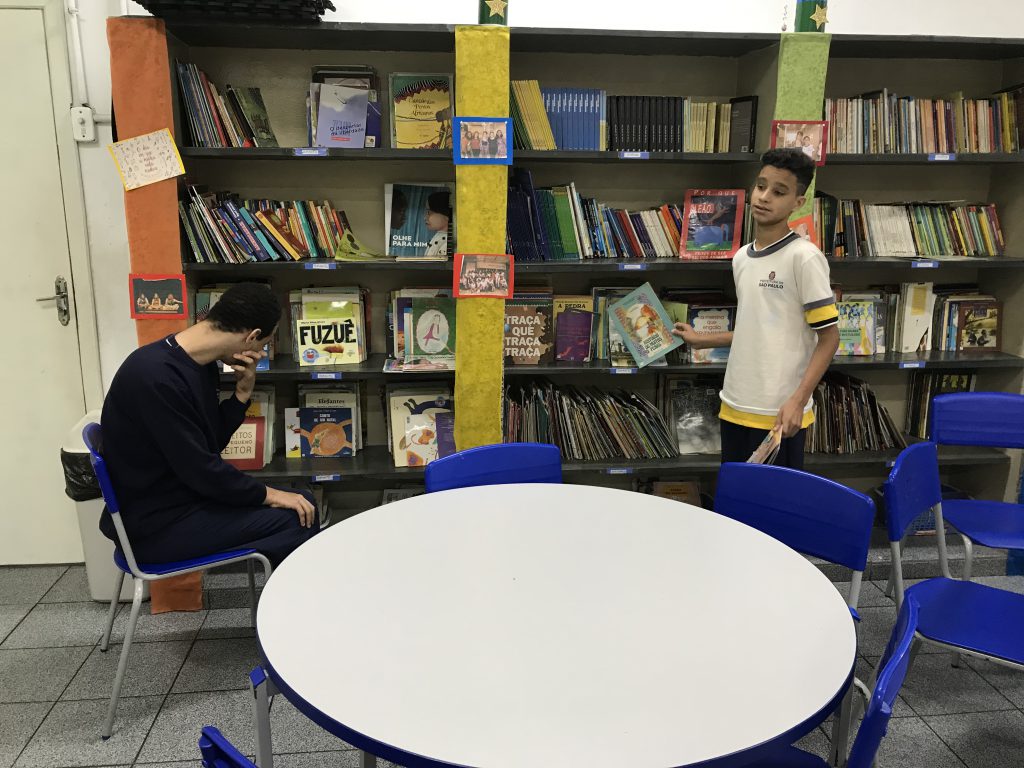 Nathan e Jonathan escolhem livros em frente à uma das estantes. Nathan está sentado à esquerda, enquanto Jonathan, à direita, está de pé. Fim da descrição.