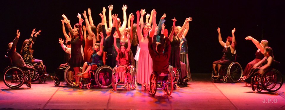 Grupo de dançarinos realiza apresentação artística em palco levantando as mãos. No grupo, há cadeirantes e pessoas sem deficiência. Fim da descrição.