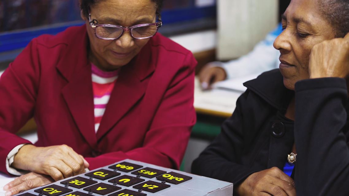 Duas senhoras negras sentadas em carteiras escolares interagem com o material pedagógico acessível Caixa silábica. Fim da descrição.