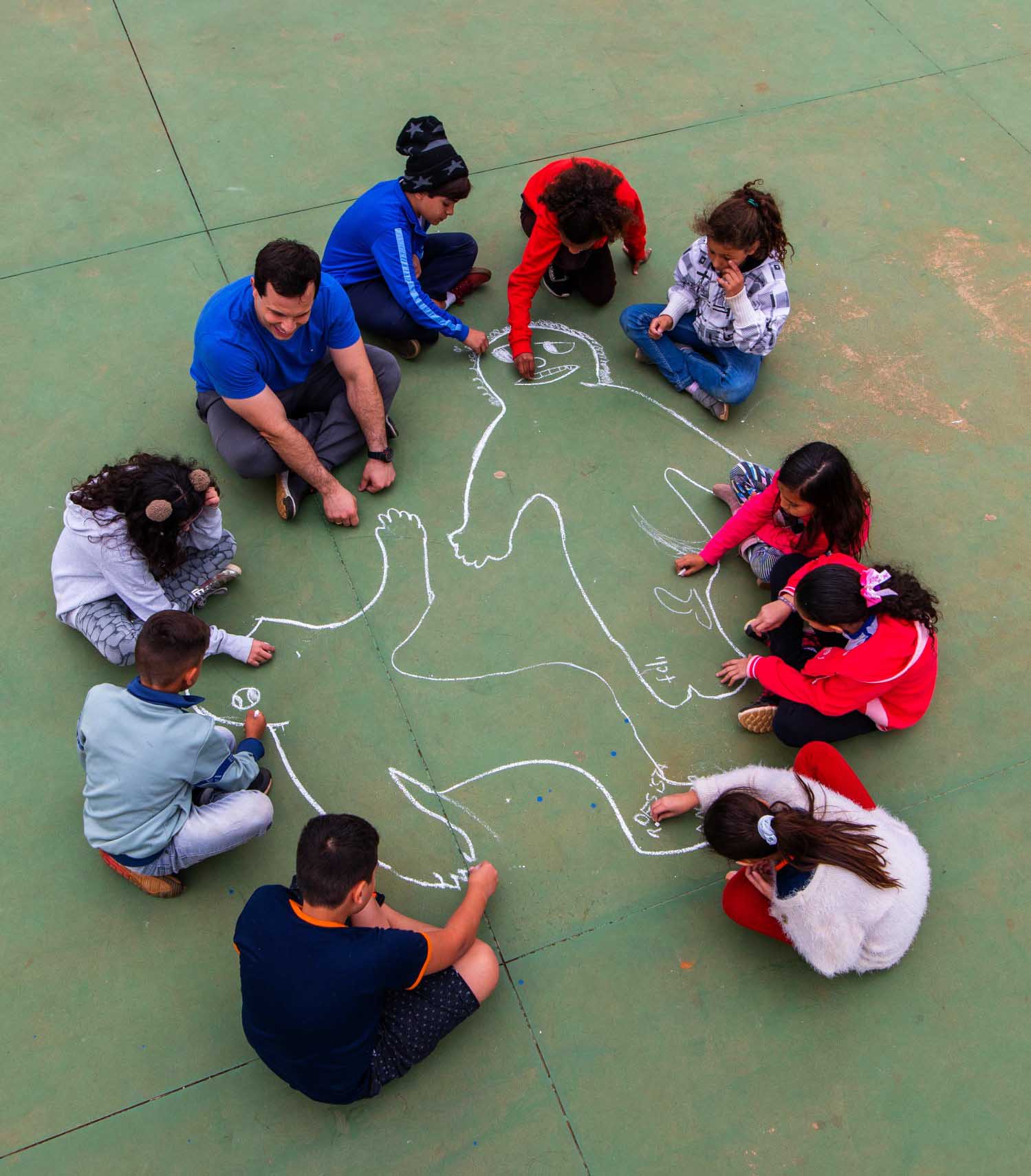 Imagem aérea do professor Luiz Gustavo e sua turma desenhando corpos no chão da quadra poliesportiva, usando giz. Fim da descrição.