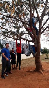O professor Luiz Gustavo posa para foto com estudantes ao lado de árvore. Os alunos estão escalando a árvore, pendurados no tronco. Fim da descrição.