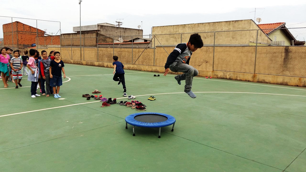 Em quadra poliesportiva, estudantes fazem fila para pular em trampolim. Um aluno encontra-se no ar depois de ter pulado. Fim da descrição.