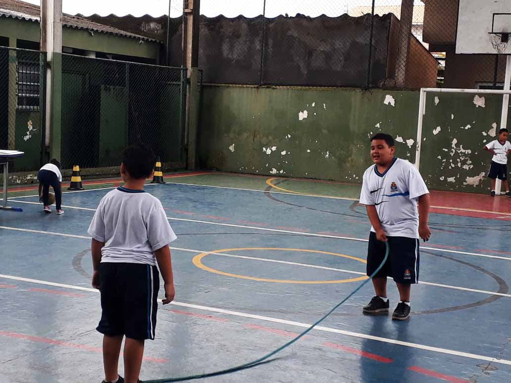 Em quadra poliesportiva de escola, dois estudantes aparecem pulando corda. Um deles está segurando a corda com a mão direita. Ambos usam uniforme escolar. Fim da descrição. 