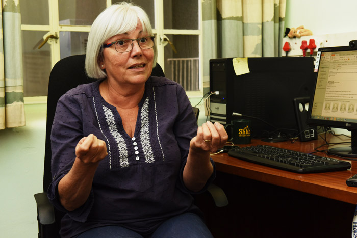 Em escritório, professora Judith Mckenzie fala enquanto gesticula com as mãos. Ela está sentada em cadeira ao lado de mesa de computador.